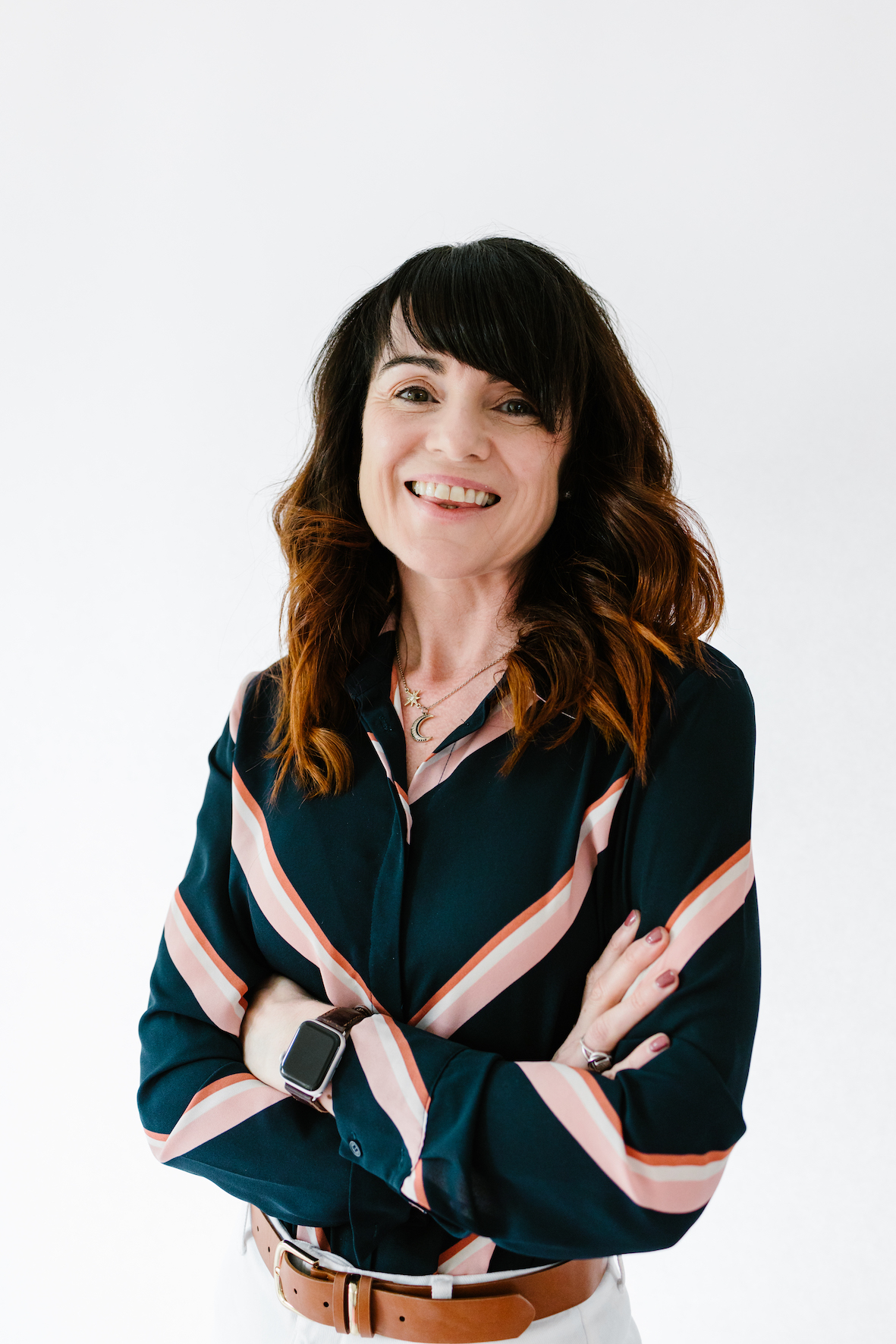 Maura is smiling at the camera. She is wearing a black blouse with pink stripes, and has long brown hair.